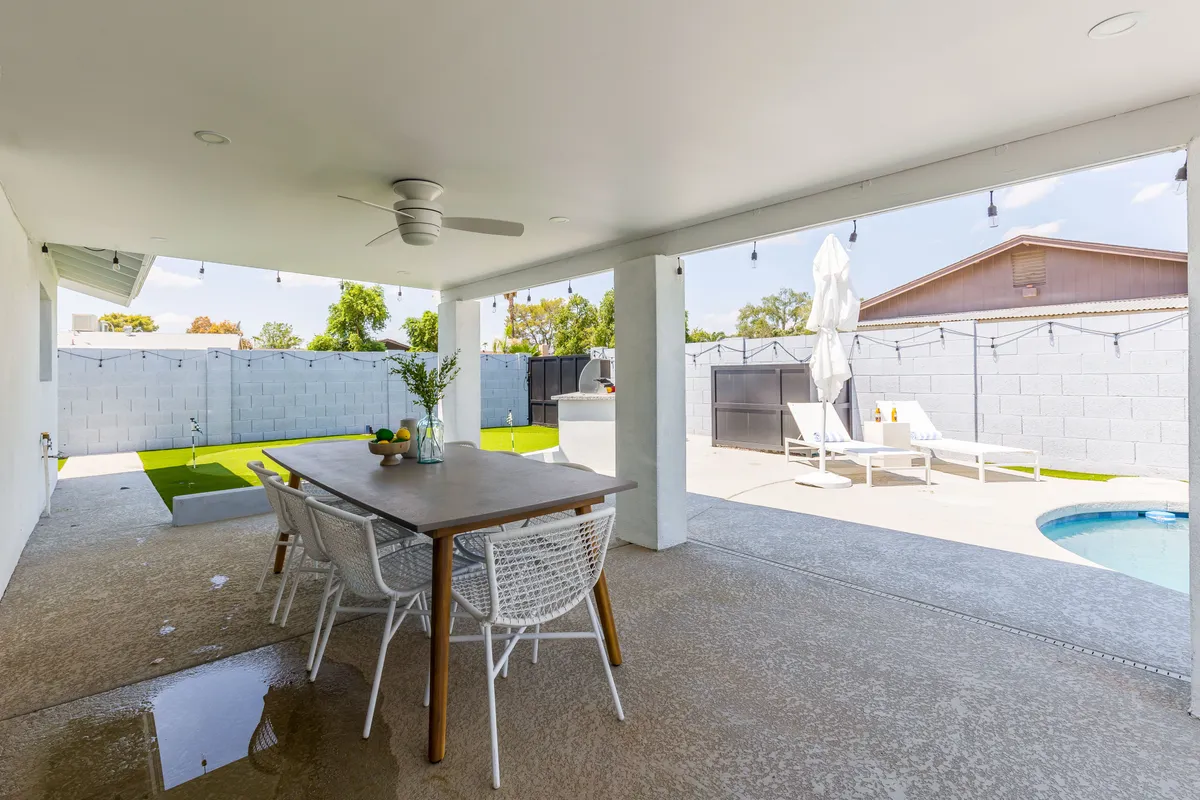 This photo presents an outdoor dining table shaded by an overhang, situated next to a shimmering pool and surrounded by lounge chairs. The setup suggests a seamless transition from swimming to alfresco dining and relaxation, with an outdoor ceiling fan adding comfort for all-day enjoyment.