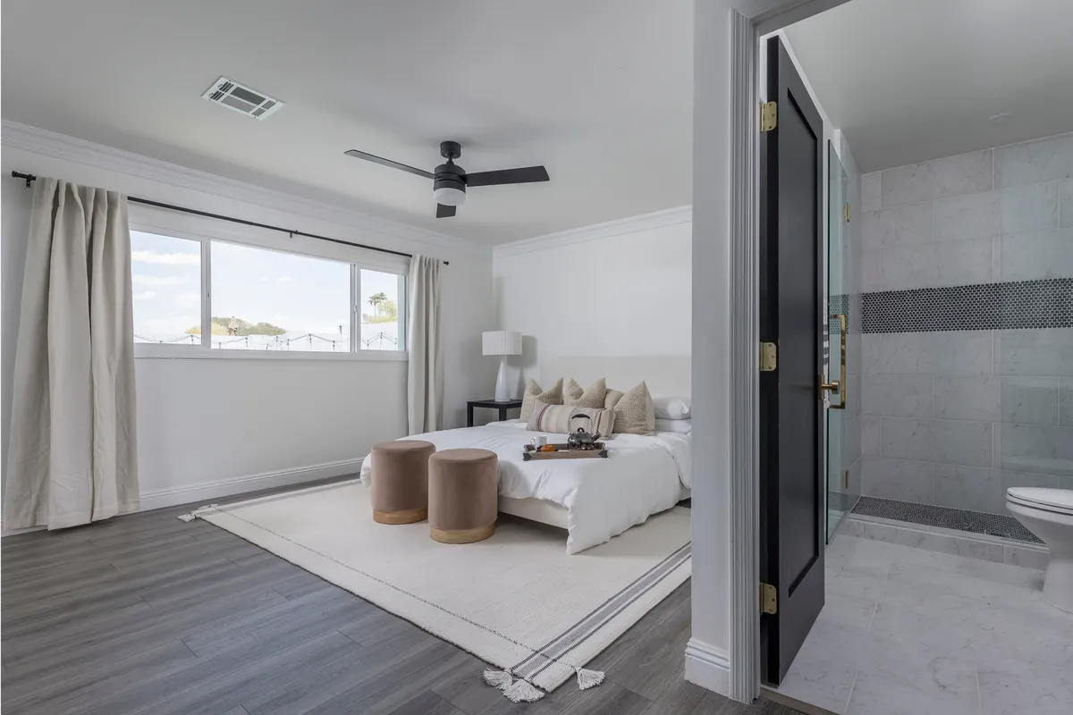 A tranquil bedroom featuring a plush bed with decorative pillows, two round stools at the foot, a large window that bathes the room in natural light, and an open doorway revealing a glimpse of a modern en-suite bathroom. The atmosphere is serene, perfect for restful sleep and relaxation.