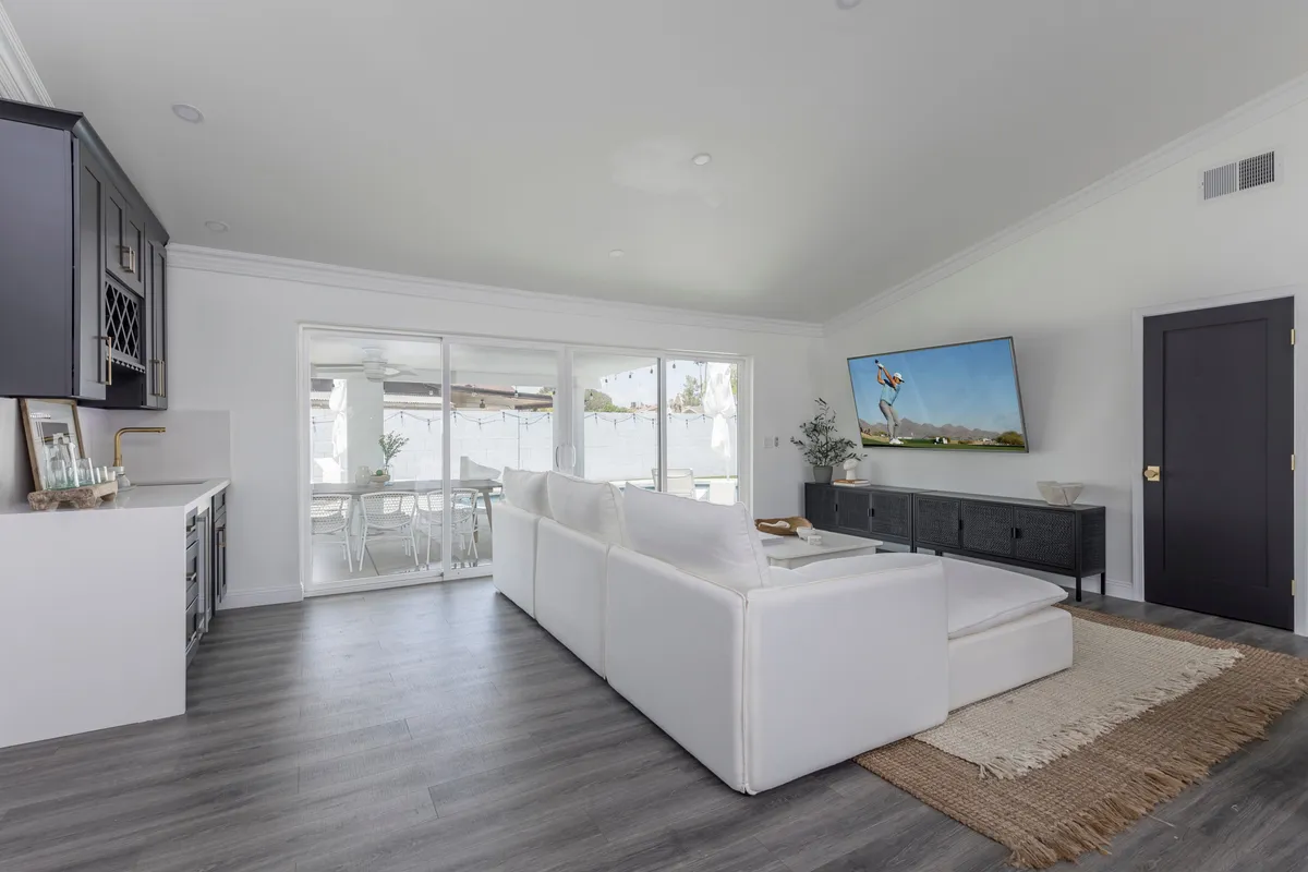 A bright, open living area featuring a plush white sectional sofa positioned to face a large smart TV. Expansive glass sliding doors allow natural light to flood the space, while the adjacent kitchen cabinetry hints at the seamless open-concept design. The décor is modern yet inviting, with thoughtful details creating a cozy, sophisticated ambiance ideal for relaxation or entertaining.