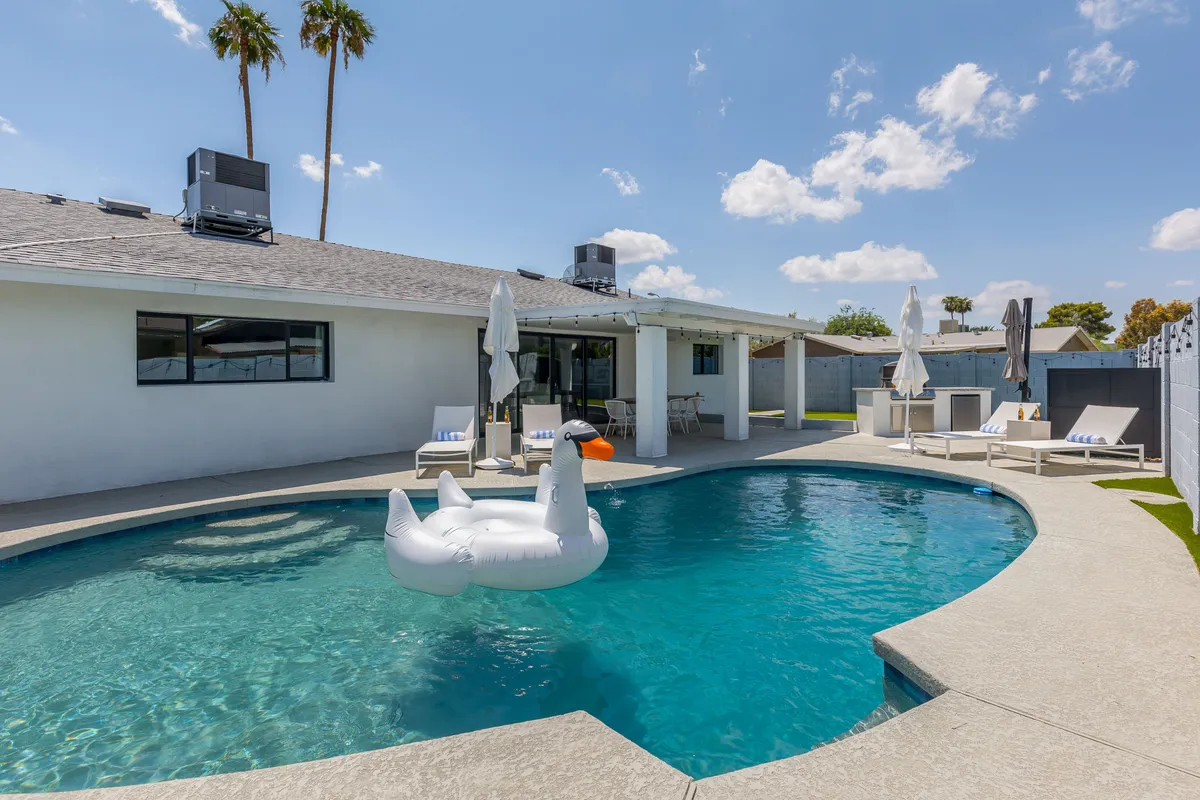 A vibrant exterior shot of a private backyard featuring a sparkling swimming pool adorned with an inflatable swan, surrounded by palm trees and comfortable lounge chairs. The inviting setup exudes a family-friendly, resort-like atmosphere perfect for relaxation and play.