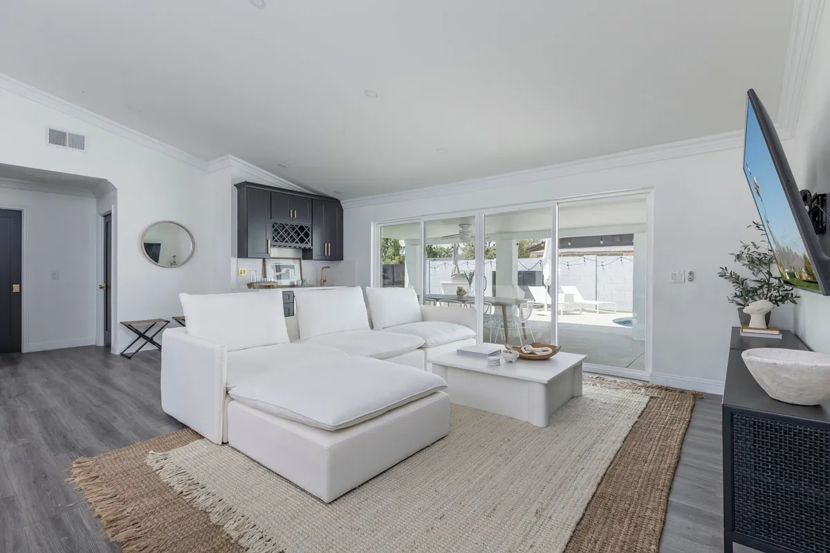 A stylish, airy living space centered on a white sectional sofa and modern coffee table, with large windows letting in streams of light. The space seamlessly connects to the kitchen area, emphasizing the open-concept design. Thoughtful, curated décor pieces add character without cluttering the room, reinforcing the balance between luxury and comfort.