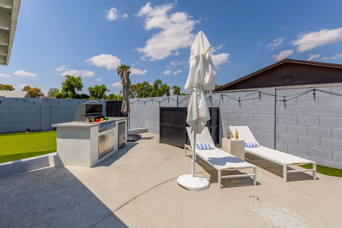 An inviting backyard featuring an outdoor kitchen with grill, lounge chairs shaded by an umbrella, and a lush green lawn. The space is designed for relaxation, socializing, and enjoying outdoor living, embodying the essence of a private retreat.